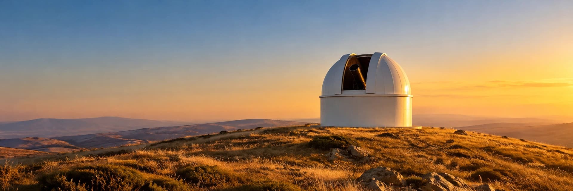 Editorial photograph at golden hour of a small white astronomical observatory standing alone on a dry grassy ridge, with scattered rocks in the foreground and hazy rolling hills behind. The dome's slit is open and the tube of a telescope is just visible inside. Low amber light from the right catches the dome's right edge, and the sky grades from warm gold at the horizon to pale blue above. No people, vehicles, lettering, logos or other buildings appear in the frame.