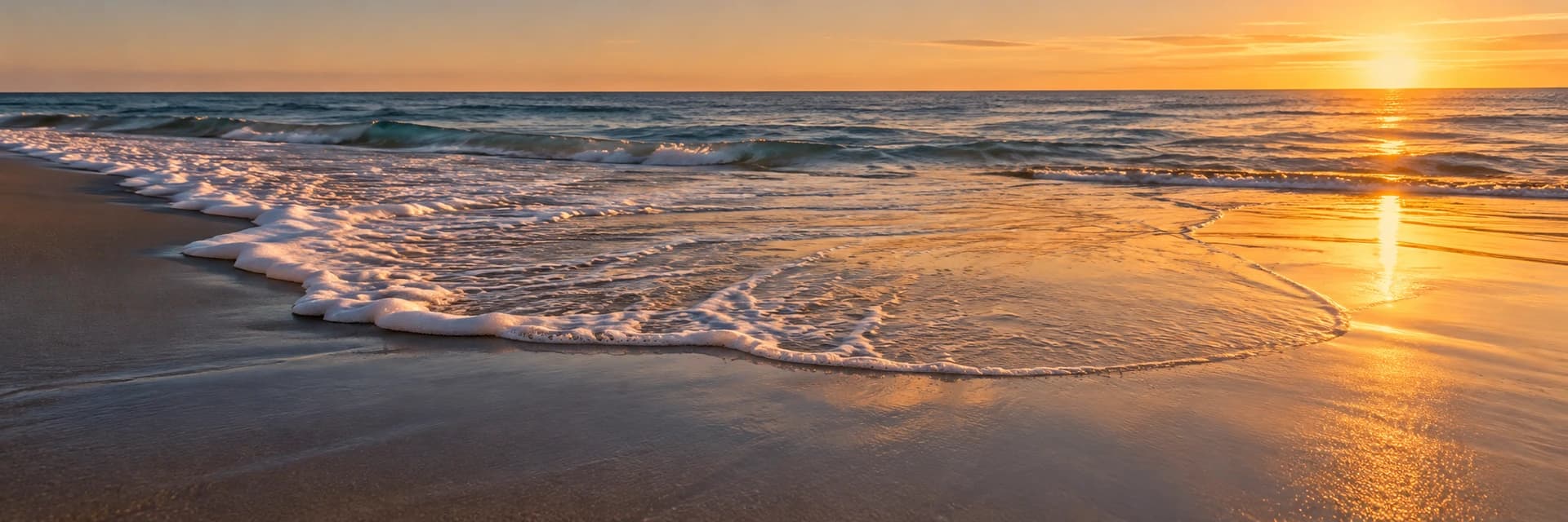 Editorial photograph at golden hour of a single spent wave running up a wide, flat sand beach. Near the center of the frame the white foam line thins into a broad, glassy sheet of water that spreads across the wet sand and flattens to a mirror-like edge. Low amber sun from the right throws long reflections across the wet sand, and a gently breaking sea sits under a saturated amber evening sky. No people, no boats, no buildings and no lettering appear anywhere in the frame.